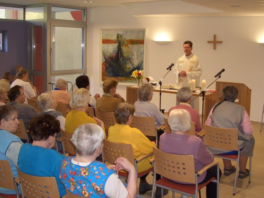 Ein Pfarrer steht beim Altar und viele BewohnerInnen sitzen auf Sesseln.